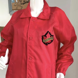 May include: A bright red windbreaker jacket featuring a pointed collar, long sleeves with elastic cuffs, and a front snap-button closure. On the left chest, there is a black embroidered patch shaped like a maple leaf with the word CANADA in yellow.