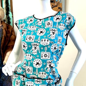 Puede incluir: Vestido turquesa sin mangas con ribete negro y un caprichoso estampado blanco de varias formas y símbolos. El vestido se exhibe en un maniquí. El vestido tiene dos bolsillos y un ribete negro.