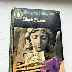 Puede incluir: Libro de bolsillo vintage "Black Plumes" de Margery Allingham. La portada presenta una imagen en escala de grises de una estatua de ángel, una mano de esqueleto y una cinta morada. El libro tiene lomo verde y está desgastado.