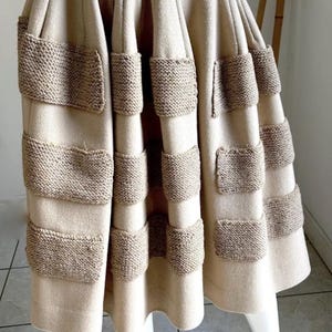 Puede incluir: Una falda beige hasta la rodilla con un diseño plisado estructurado. La falda está acentuada con filas horizontales de parches rectangulares de punto marrón texturizado aplicados en la parte delantera, creando un aspecto en capas.