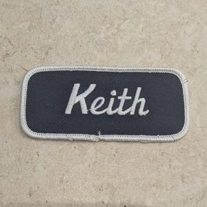 May include: A rectangular name patch with rounded corners. The patch is dark blue with a white embroidered border and the name "Keith" in white script. The patch is on a light brown, textured surface.