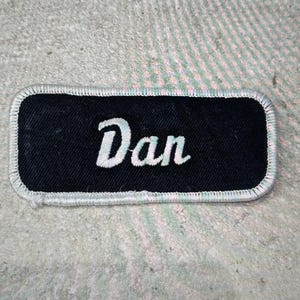 May include: A rectangular embroidered patch with a black fabric background and a white stitched border. The name Dan is embroidered in the center in a white cursive script font. The patch has rounded corners and a clean, finished edge.