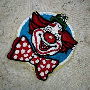 May include: An embroidered patch featuring a clown with red hair, a green hat with a white flower, and a large red bow tie with white polka dots. The clown has a white face, red nose, and wide smile, set against a circular blue background.