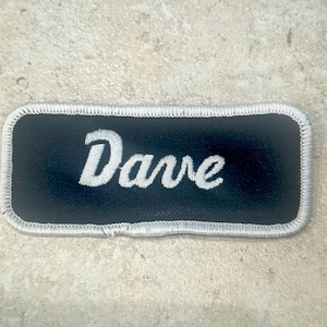 May include: A rectangular embroidered patch with a black background and a white border. The name Dave is stitched in the center in a white, cursive script font. The patch has rounded corners and a textured fabric appearance throughout the design.