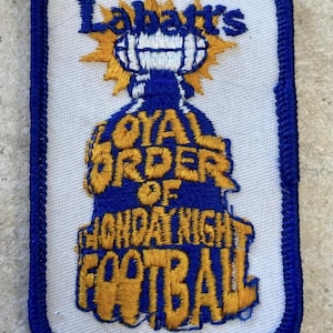 May include: A rectangular embroidered patch with a white background and a blue border. It features the text Labatt's, Loyal Order of Monday Night Football in yellow and blue thread, with a graphic of a football centred above the text.
