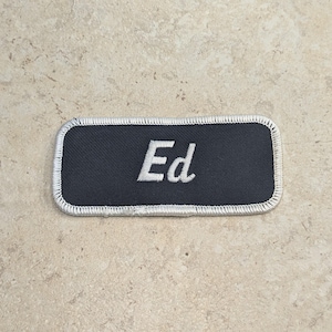 Peut inclure: Un écusson rectangulaire noir avec le nom "Ed" brodé en blanc. L'écusson a une bordure argentée et est probablement conçu pour être cousu sur des vêtements ou des accessoires. L'écusson est sur une surface de ton neutre.