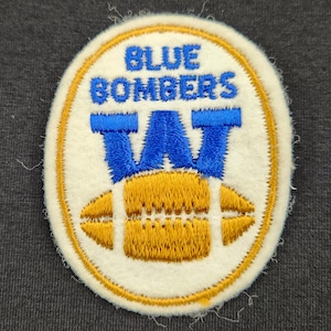 May include: An oval-shaped embroidered patch with a white background and a gold border. It features the blue text BLUE BOMBERS above a large blue letter W, which sits over a gold-colored football graphic with detailed stitching.