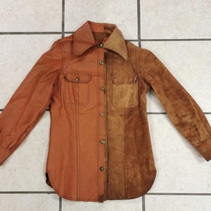 May include: A long-sleeve shirt featuring a split design with one side made of smooth tan leather and the other side made of textured brown suede. The shirt includes a pointed collar, a front button closure, and two chest pockets with flaps.