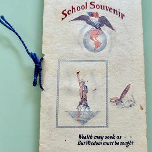 Puede incluir: Libro de recuerdos escolares vintage con una cuerda azul. La portada presenta un águila con una bandera estadounidense, un globo terráqueo y la Estatua de la Libertad. El texto dice "School Souvenir" y "Wealth may seek us - But Wisdom must be sought."