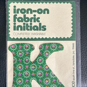 May include: An iron-on fabric initial 'K' in a clear plastic packet. The initial is green with a floral pattern of pink and purple flowers. The packaging reads 'iron-on fabric initials' and 'completely washable'.
