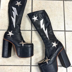 Puede incluir: Un par de botas negras de plataforma con detalles de relámpagos y estrellas plateadas. Las botas tienen un tacón grueso y una suela de plataforma. Botas altas hasta la rodilla de material similar al cuero.