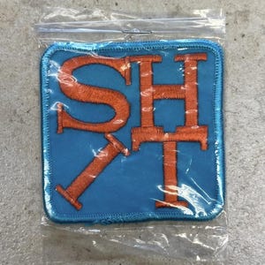 Könnte beinhalten: Ein quadratischer Stoffaufnäher mit abgerundeten Ecken, der einen hellblauen Hintergrund und gestickte orangefarbene Buchstaben mit der Aufschrift SHIT aufweist. Der Aufnäher befindet sich in einer durchsichtigen, versiegelten Plastiktüte.