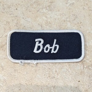 May include: A rectangular name patch with the name "Bob" embroidered in white script on a navy blue background. The patch has a white border and is likely for a uniform or workwear.