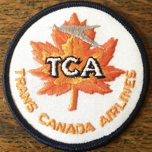 Puede incluir: Un parche bordado circular con fondo blanco y borde azul oscuro. El centro presenta una hoja de arce naranja con el texto negro TCA y una silueta de avión plateada. El texto TRANS CANADA AIRLINES está bordado en color naranja.