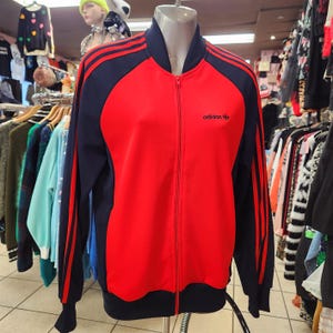 Puede incluir: Chaqueta deportiva roja y azul marino con cremallera y el logo de Adidas. La chaqueta tiene rayas azules marino en las mangas y cuello y puños azules marino. La chaqueta se exhibe en un maniquí.