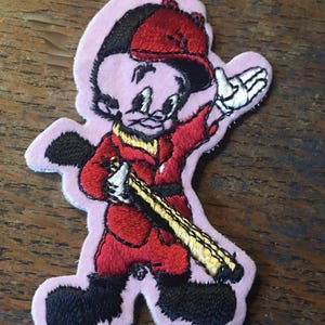 May include: An embroidered fabric patch featuring a cartoon character wearing a red cap, a red jacket, and black boots. The character holds a long, yellow-toned object across the body and has one hand raised. The patch has a light pink border.