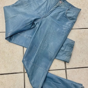 May include: Light blue leather pants with a classic five-pocket design. The pants have a button and zipper closure and belt loops. The leather has a slightly distressed appearance. The pants are laid flat on a tiled surface.