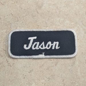 May include: A rectangular name patch with the name "Jason" embroidered in white script on a navy blue background. The patch has a white stitched border.