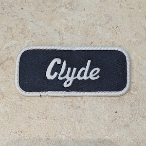 May include: A rectangular patch with the name "Clyde" embroidered in white script on a navy blue background. The patch has a white stitched border and is set against a neutral, speckled surface.
