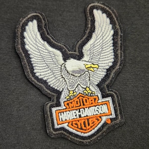 May include: An embroidered patch featuring a white eagle with spread wings, a yellow beak, and yellow talons. Below the eagle is an orange and white shield-shaped emblem with the text Harley-Davidson Cycles in black lettering on a white background.