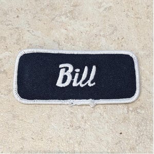 Op de afbeelding: Een rechthoekige marineblauwe naamvlek met de naam "Bill" in witte letters geborduurd. De vlek heeft een wit gestikte rand en is aan de onderkant licht gerafeld.