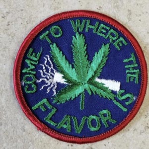 Puede incluir: Un parche bordado circular con fondo azul oscuro y borde rojo. Presenta una hoja de cannabis verde, un cigarrillo blanco con humo blanco y el texto bordado en verde "COME TO WHERE THE FLAVOR IS" rodeando la hoja.
