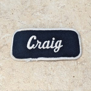 May include: A rectangular navy blue name patch with the name "Craig" embroidered in white script. The patch has a white stitched border and is likely for a uniform or workwear.
