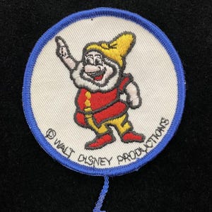 May include: A circular embroidered patch featuring a cartoon character with a yellow hat, red tunic, and yellow pants. The character is waving one hand. The patch has a blue border and the text "© WALT DISNEY PRODUCTIONS" printed along the bottom edge.
