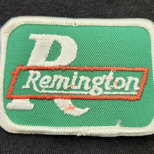 May include: A rectangular embroidered patch featuring a green background with a white border. A large white letter R is centred behind a horizontal orange rectangular bar containing the white text Remington in a cursive script font.