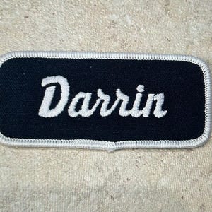 May include: A rectangular embroidered patch with rounded corners featuring a black background and a white border. The name Darrin is stitched in white cursive script across the center of the patch. The design is clean and simple with clear lettering.