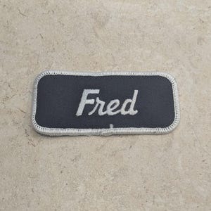 May include: A rectangular name patch with the name "Fred" embroidered in white script on a dark gray background. The patch has a white border and is likely for a uniform or clothing item.
