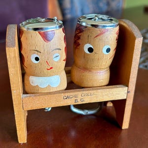 May include: Wooden salt and pepper shakers in the shape of cartoon figures, with painted faces and metal tops. The shakers are set in a wooden holder, with the text "CACHE CREEK B.C." on the front.
