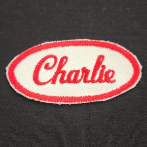 May include: An oval-shaped embroidered patch featuring a white background with a red border. The name Charlie is stitched in red cursive script across the centre of the patch. The embroidery has a visible textured thread pattern throughout.