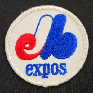 Puede incluir: Un parche bordado circular con fondo blanco y un logotipo estilizado en azul y rojo. El logotipo consiste en una e minúscula roja y una b minúscula azul entrelazadas. Debajo del logotipo, la palabra expos está bordada en letras azules.