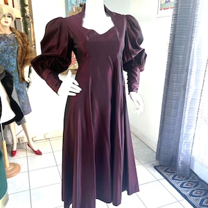 May include: A burgundy vintage dress with a high neckline and long sleeves. The dress has a fitted bodice and a flared skirt. The sleeves have a puffed design at the shoulders. The dress is displayed on a mannequin.