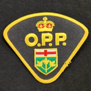 Peut inclure: Un écusson brodé triangulaire avec un fond gris foncé et une bordure jaune épaisse. Il présente une couronne jaune au-dessus du texte jaune O.P.P. et un écusson affichant une croix rouge sur fond blanc au-dessus de trois feuilles d'érable jaunes.