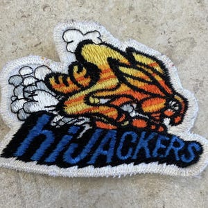 May include: An embroidered fabric patch featuring a cartoon character with orange and yellow fur running quickly, leaving a trail of white smoke behind. The word "hiJACKERS" is stitched in bold blue letters with a black outline below the character.
