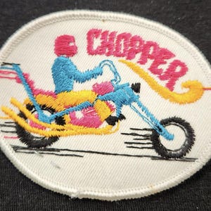 May include: An oval-shaped embroidered patch on a white background featuring a stylized figure in a pink helmet riding a blue and yellow motorcycle. The word CHOPPER is stitched in pink above the motorcycle, with a yellow flame design trailing behind.