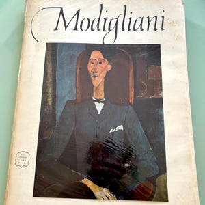 May include: A vintage book titled "Modigliani" featuring a portrait of a man in a dark suit. The cover has the text "16 Beautiful Full Color Prints" and an "An Art Book" emblem.
