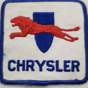 Puede incluir: Un parche bordado cuadrado con fondo blanco y borde azul. Presenta un león rojo saltando sobre un escudo azul. La palabra CHRYSLER está bordada en letras mayúsculas azules en la parte inferior del parche, centrada debajo del escudo.