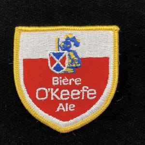 May include: A shield-shaped embroidered patch with a yellow border. The top half is white and the bottom half is red. It features a blue knight figure holding a small shield with a white cross, and the text Bière O'Keefe Ale in white.