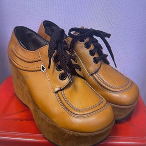 Pode incluir: Um par de sapatos de plataforma em couro castanho-claro com atacadores pretos, ilhós pretos e uma sola de cunha grossa com textura de camurça castanha. Os sapatos têm um design de biqueira redonda com costuras pretas visíveis nas laterais.