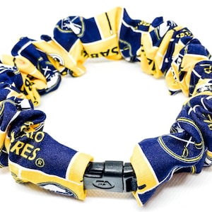 Puede incluir: Un collar de tela en azul marino y amarillo, con un patrón repetido con el texto "SABRES". El collar se asegura con un cierre de plástico negro. La tela está fruncida y agrupada, creando una apariencia texturizada.