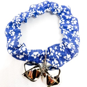 May include: Blue and white patterned cat collar with a silver clasp and two silver bells. The fabric features a repeating pattern of white paw prints and stars. The collar is designed to be stretchy.