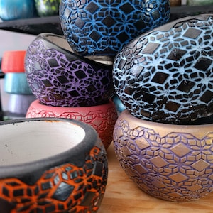 May include: A selection of decorative ceramic pots in shades of blue, purple, orange, and gold. Each pot has a geometric diamond pattern and a black rim. The pots are stacked, showcasing their contrasting colours and textures.