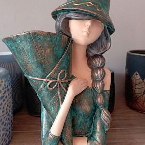 Op de afbeelding: Een decoratieve plantenbak in de vorm van een vrouw met een hoed en een gestileerd boeket. De plantenbak heeft een teal en bronzen afwerking. Het haar van de vrouw is gevlochten en haar huid is een lichte tint. Andere plantenbakken zijn zichtbaar op de achtergrond.