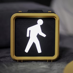 May include: A yellow pedestrian crossing signal with a white walking person icon illuminated against a black background.