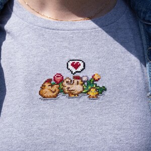 Op de afbeelding: Een grijs t-shirt met een gepixeld geborduurd ontwerp van twee kippen met een hart erboven. De kippen zijn omringd door bloemen en groen.