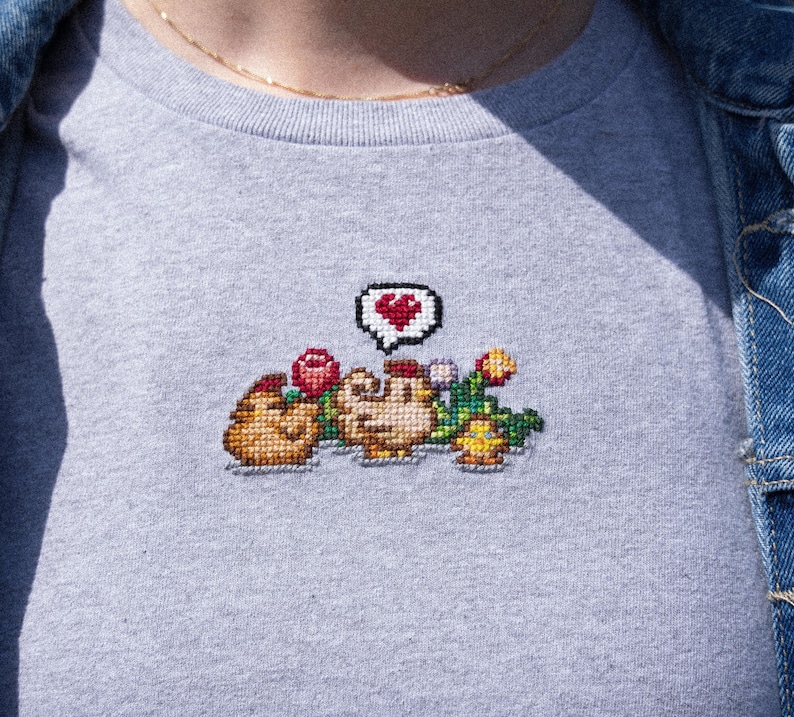 Op de afbeelding: Een grijs t-shirt met een gepixeld geborduurd ontwerp van twee kippen met een hart erboven. De kippen zijn omringd door bloemen en groen.