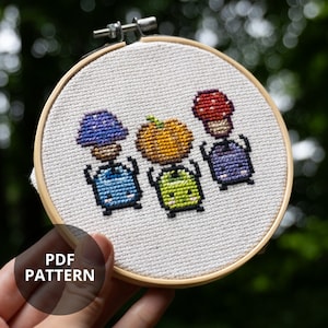 May include: A cross-stitch embroidery in a wooden hoop shows three pixelated mushroom characters with blue, orange, and red caps. The characters have blue, green, and purple bodies. The text "PDF PATTERN" is in a black circle.